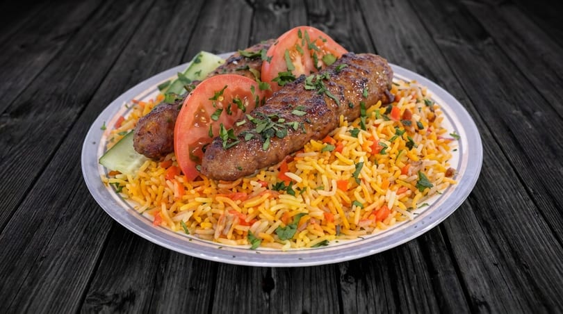 Grilled kofta kebabs on a bed of yellow rice with fresh tomatoes and cucumber slices.