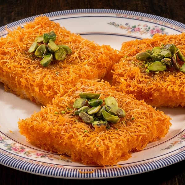 Orange shredded pastry dessert topped with green pistachios on a decorative white and blue plate.