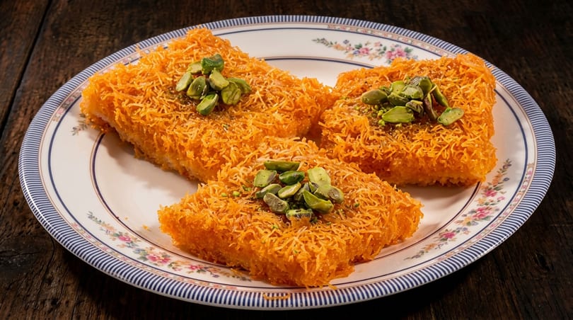 Orange shredded pastry dessert topped with green pistachios on a decorative white and blue plate.