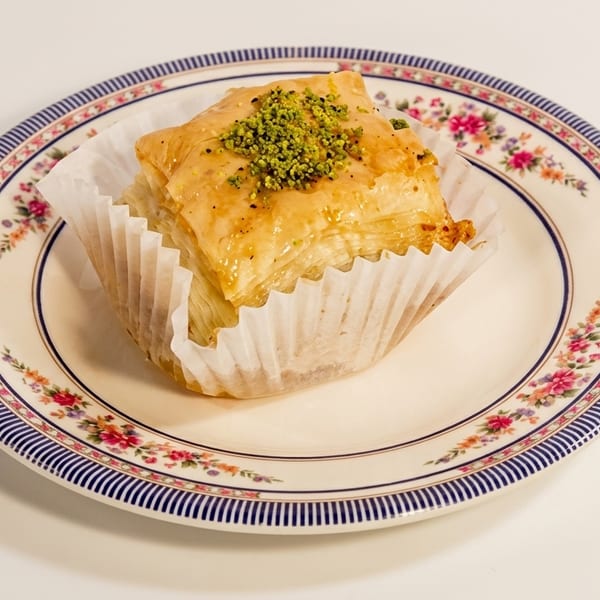 Golden baklava with pistachios in a paper wrapper on a decorative floral plate.
