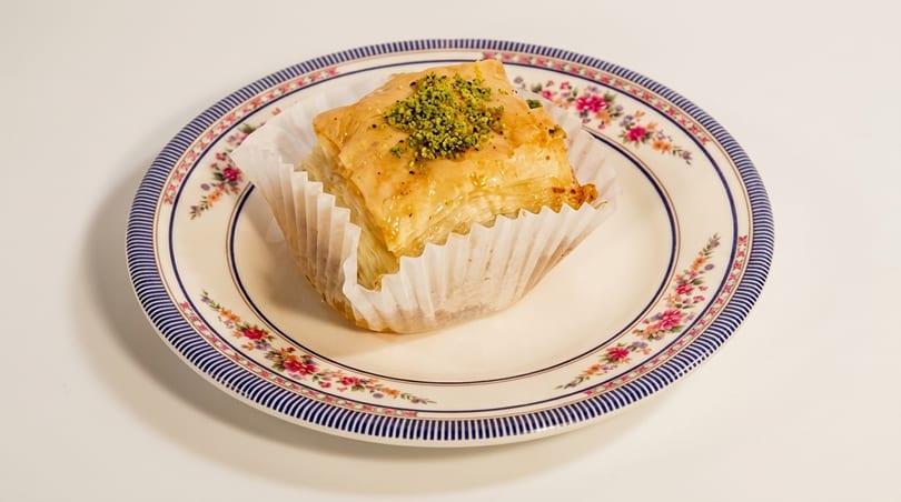 Golden baklava with pistachios in a paper wrapper on a decorative floral plate.